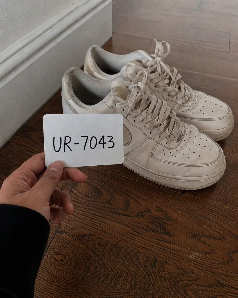 A hand holding a white card reading U R dash 7 0 4 3 beside a pair of worn white trainers on a wooden floor at home.